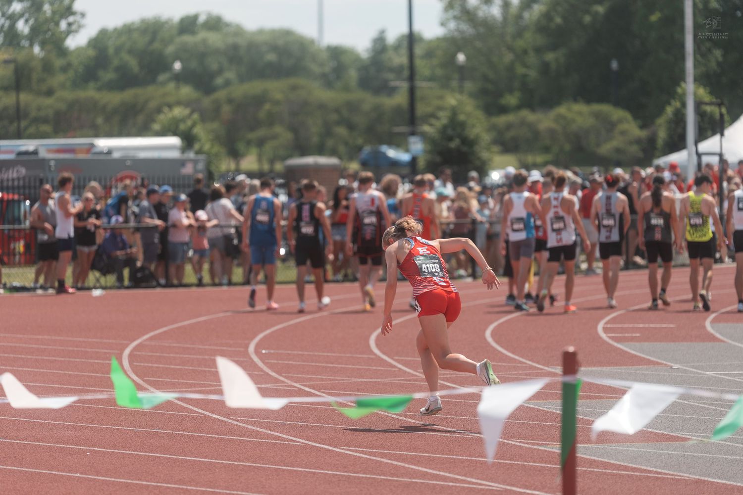 Athlete in red races around track curve at a crowded outdoor track and field competition.