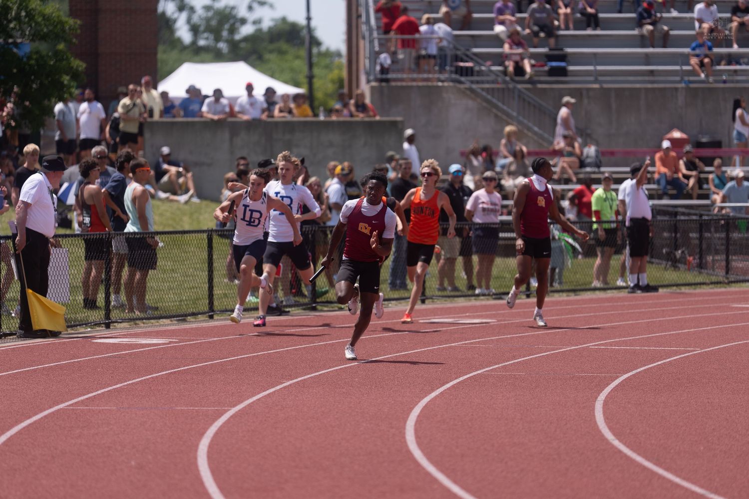 A sprinter in red leads competitors down the track at a crowded outdoor track and field event.