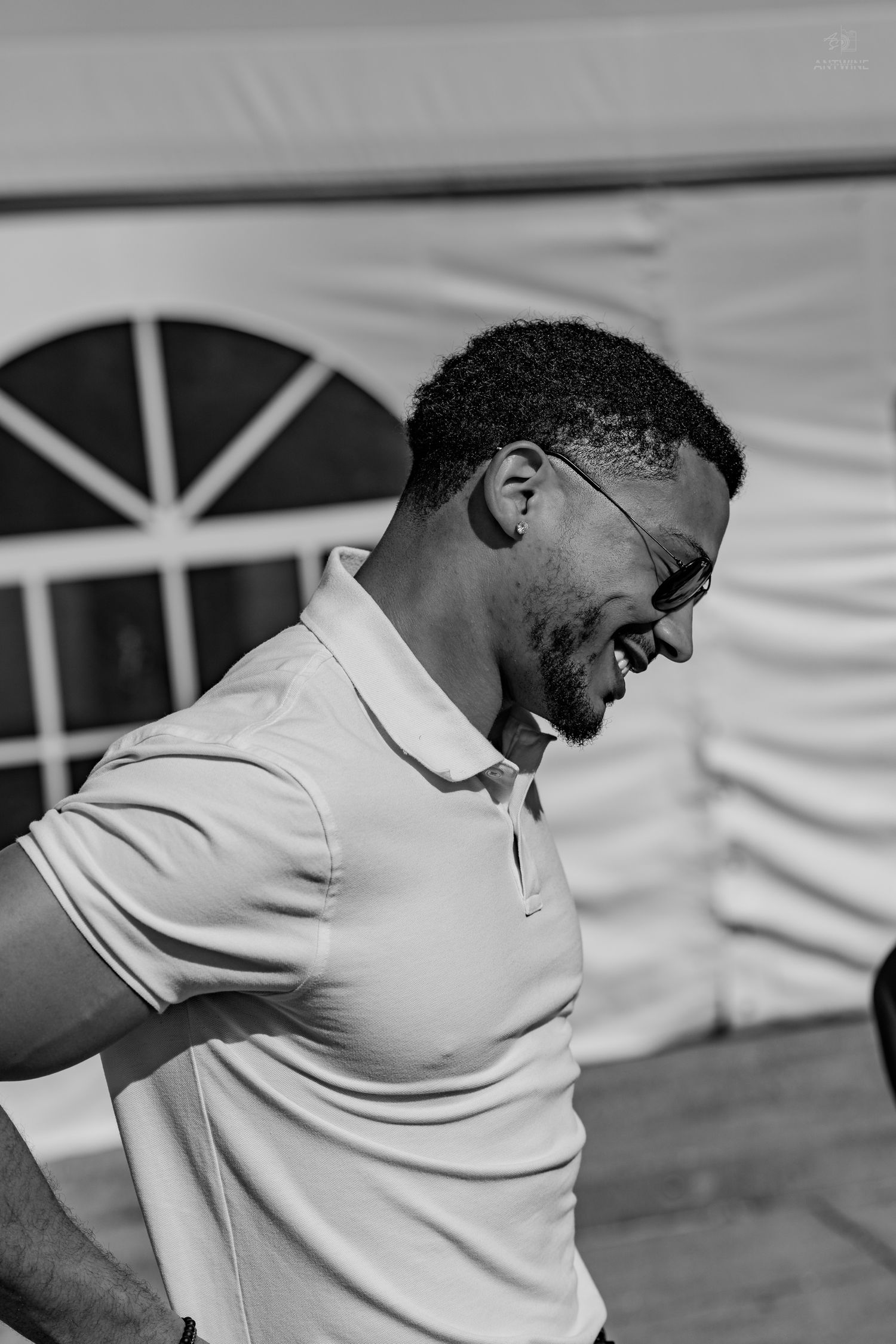 Black and white close-up of a smiling man in glasses and a polo shirt laughing near a white tent.