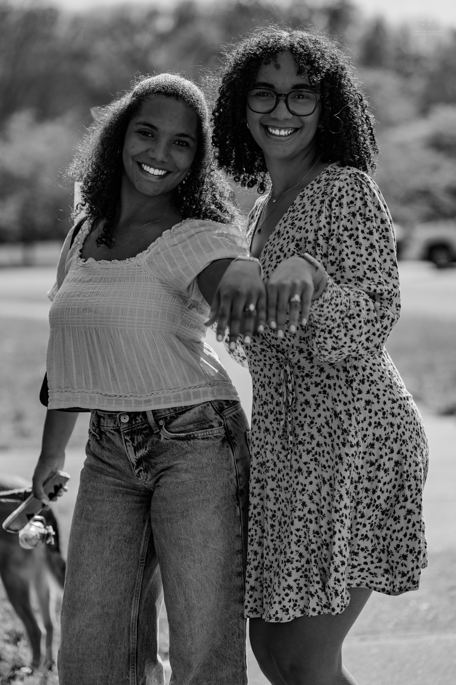 Black and white photo of two curly-haired women smiling and posing together outdoors in a sunny park setting.