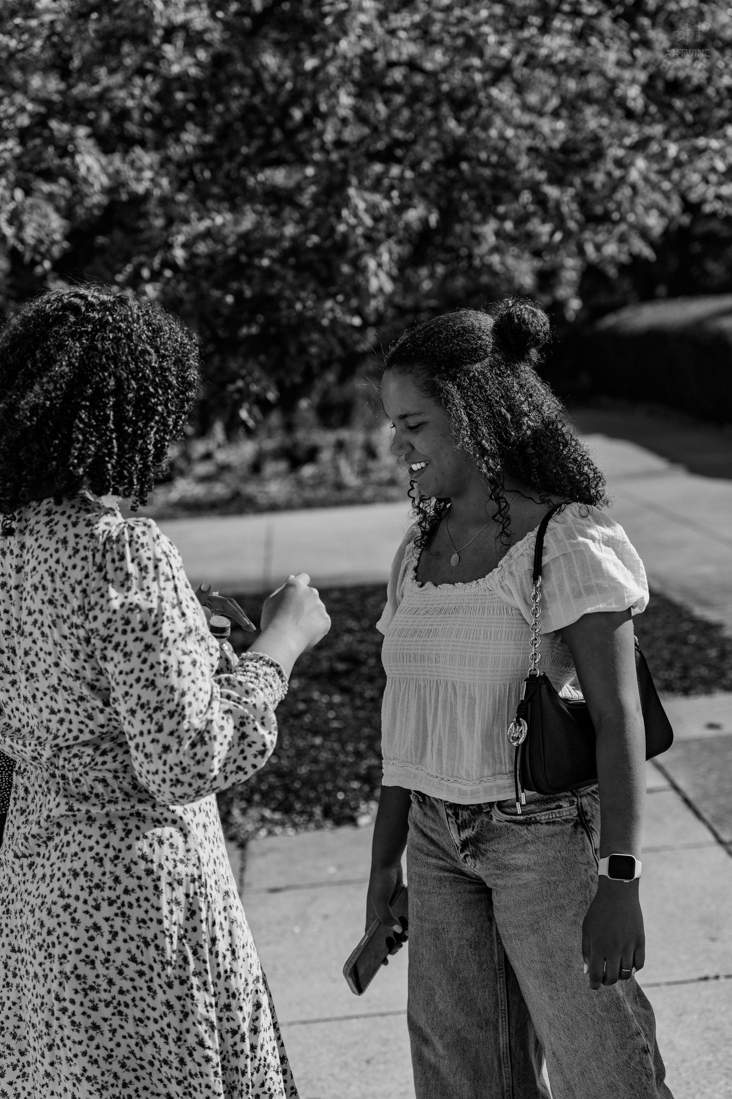 Two young Black women smile and chat outdoors on a sunny day, one wearing a white top with jeans.