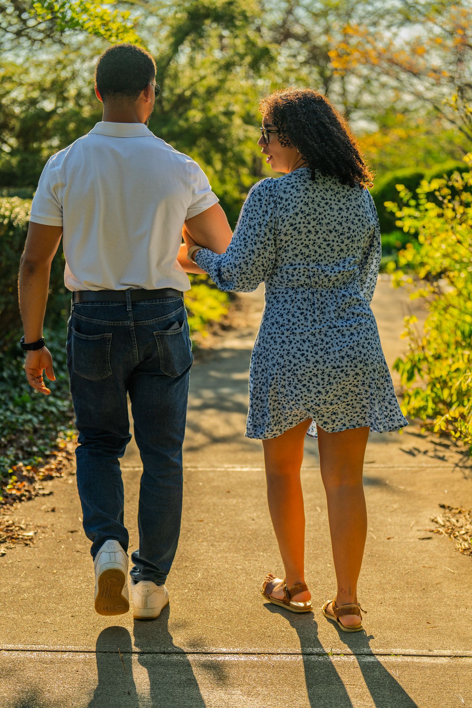 A couple holding hands while walking down a golden sunlit park path surrounded by autumn foliage.