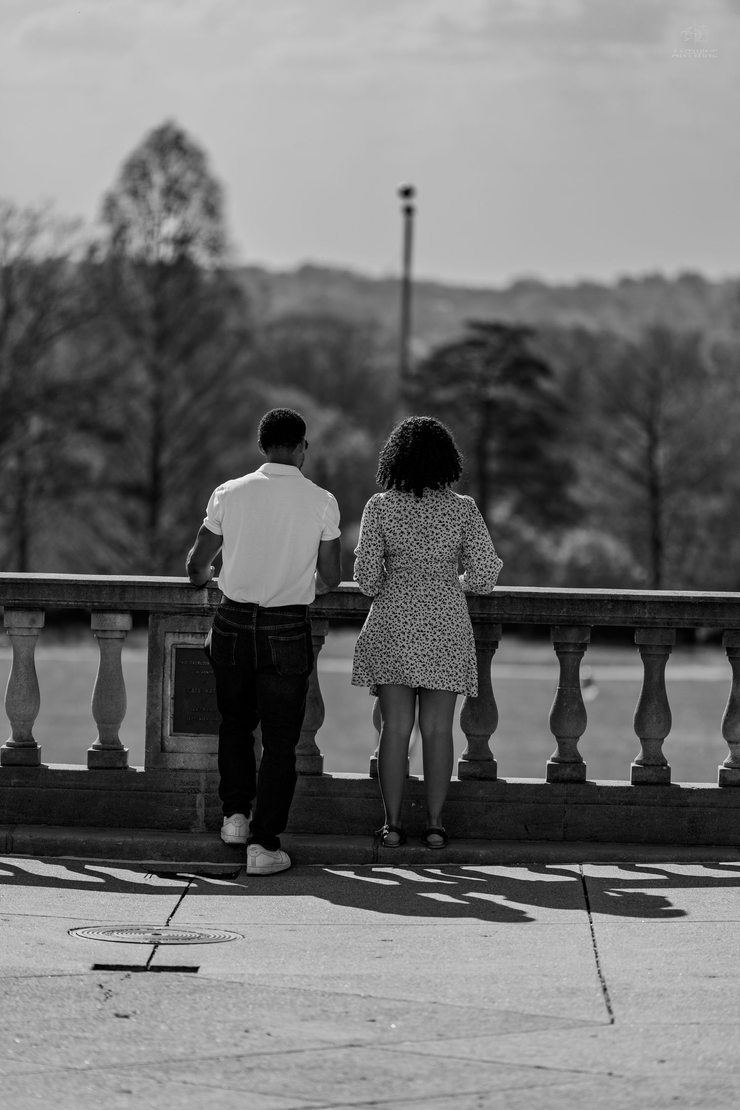 Black and white photo of a couple standing at a stone balustrade overlooking a scenic wooded landscape.