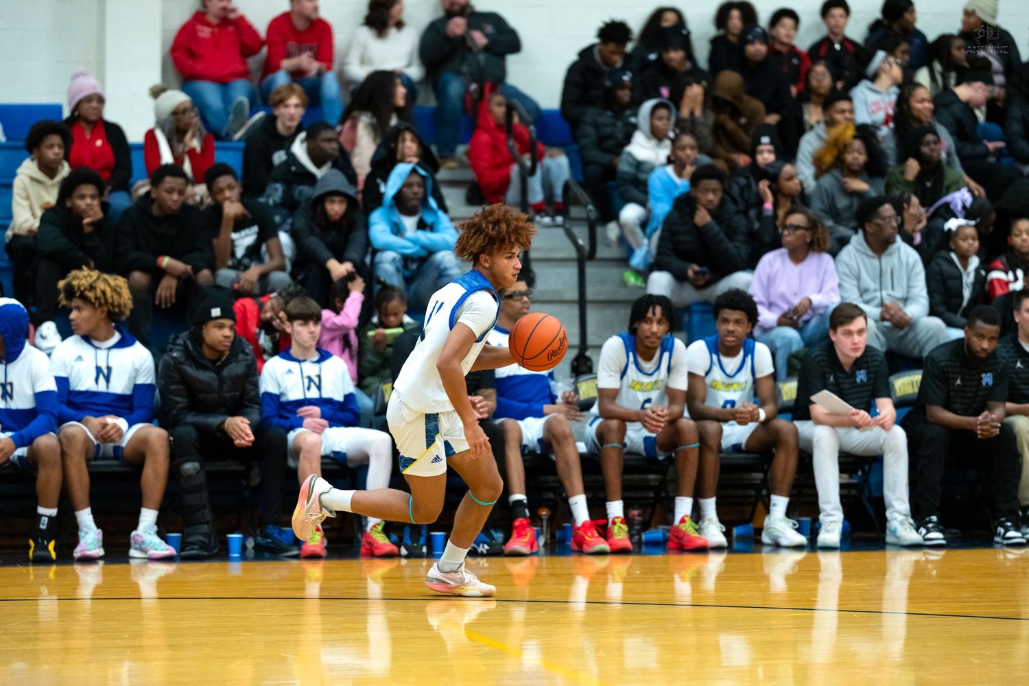 High school basketball player in white uniform dribbles ball down court in front of packed gymnasium crowd.