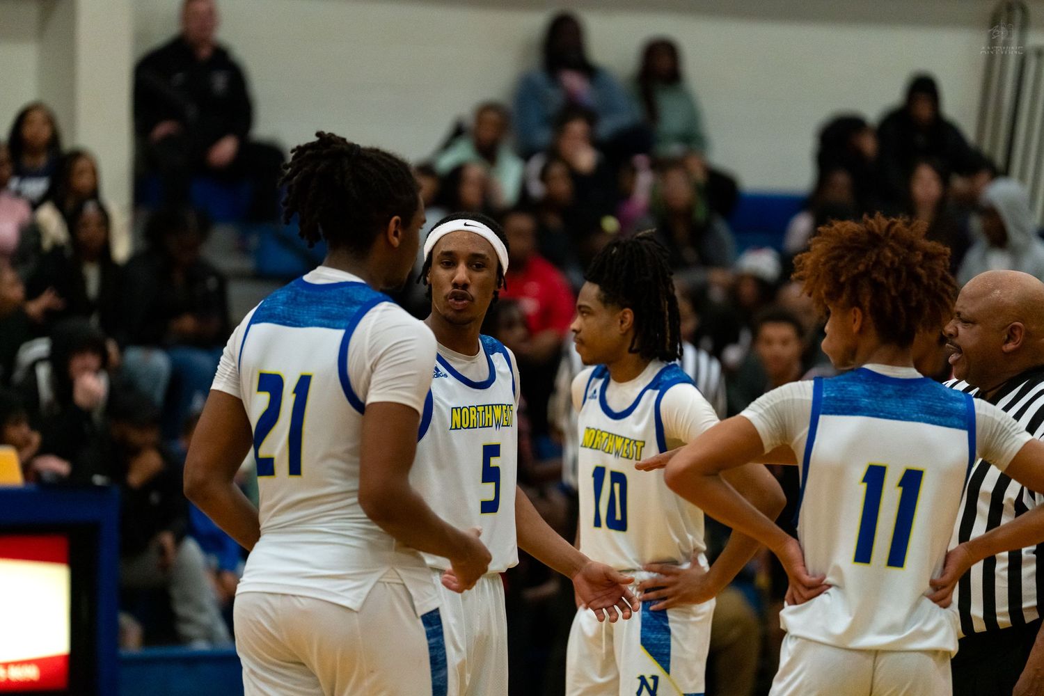 High school basketball team huddles on court wearing blue and white uniforms with numbers 21, 5, 10, and 11.