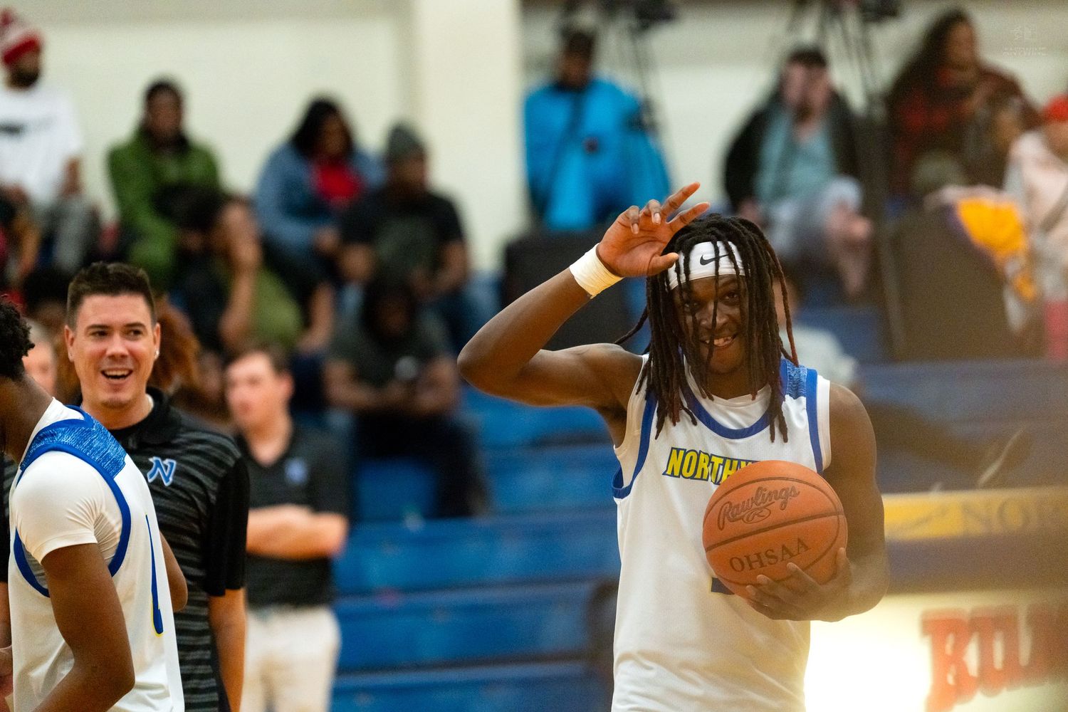 A high school basketball player holds a ball while gesturing during a game as a teammate laughs beside him.
