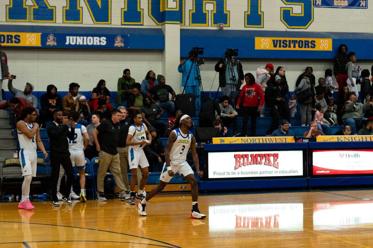 Northwest Knights players and coaches react on sideline during a home basketball game in a gymnasium with packed stands.