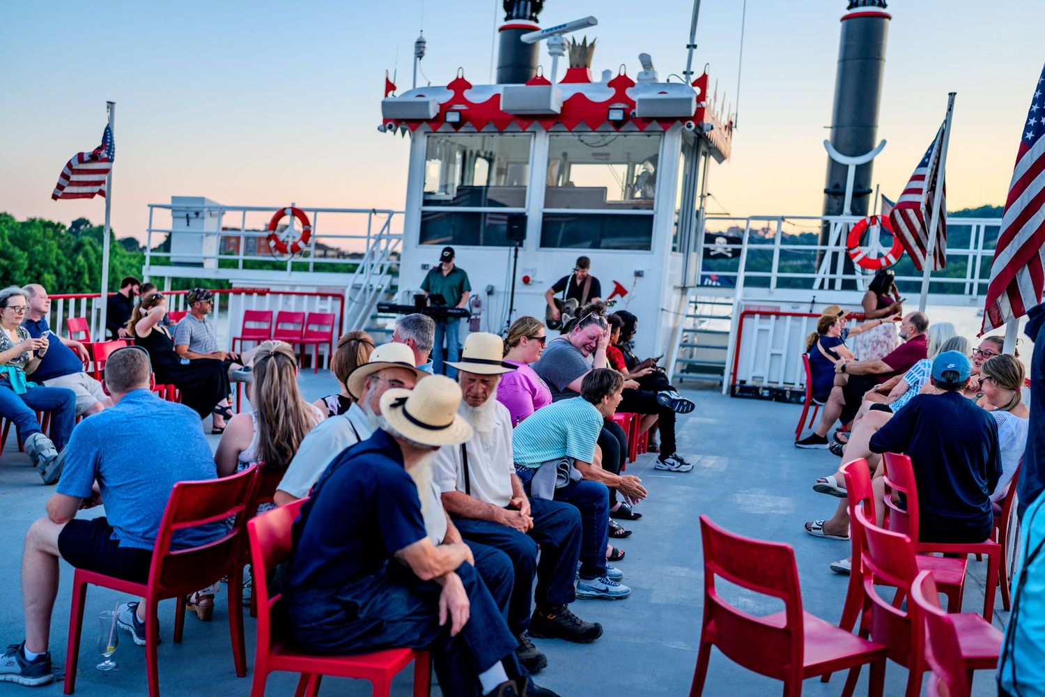Crowd of people sitting in red chairs on a boat deck enjoying a live music performance at sunset.