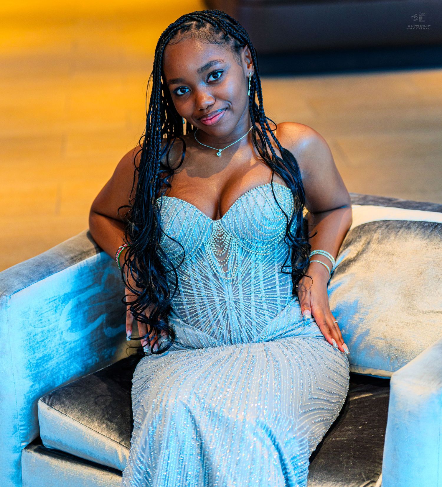 Smiling young woman in a beaded strapless silver gown with long braids seated on a velvet chair.