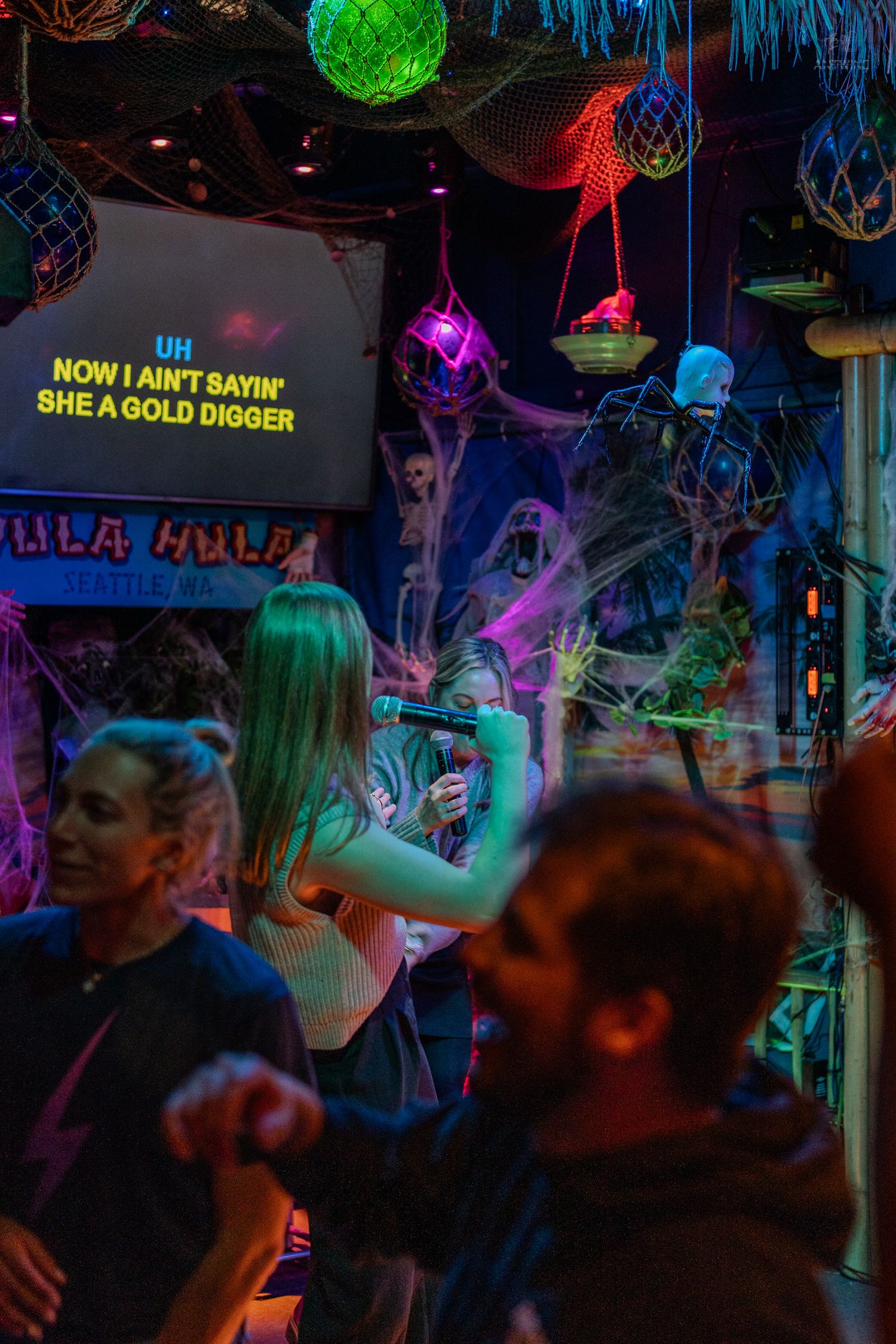 Woman singing karaoke at Hula Hula bar with neon lights and Halloween spider web decorations.