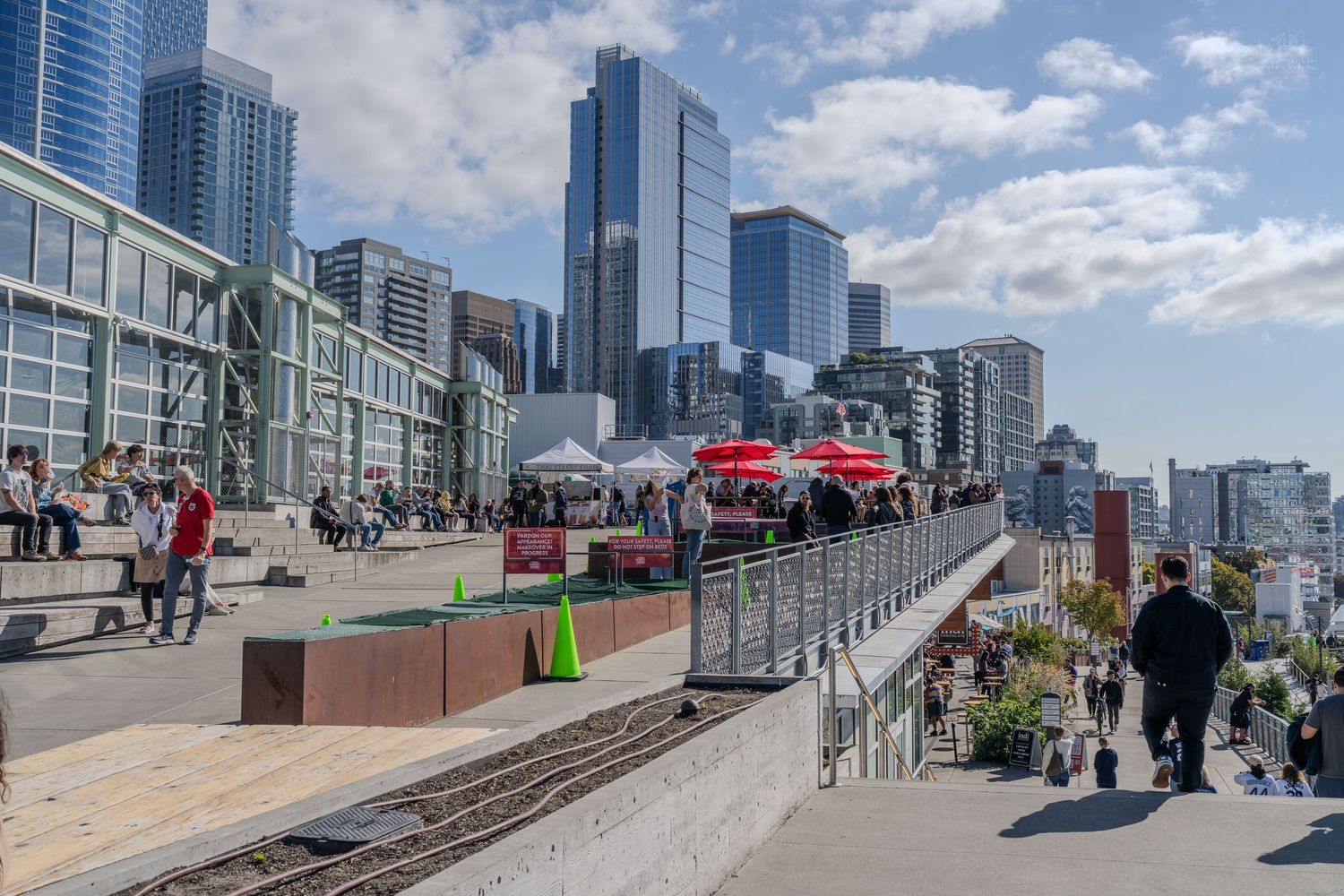 Busy waterfront promenade with city skyline, pedestrians, food trucks, and railway tracks on a sunny day.