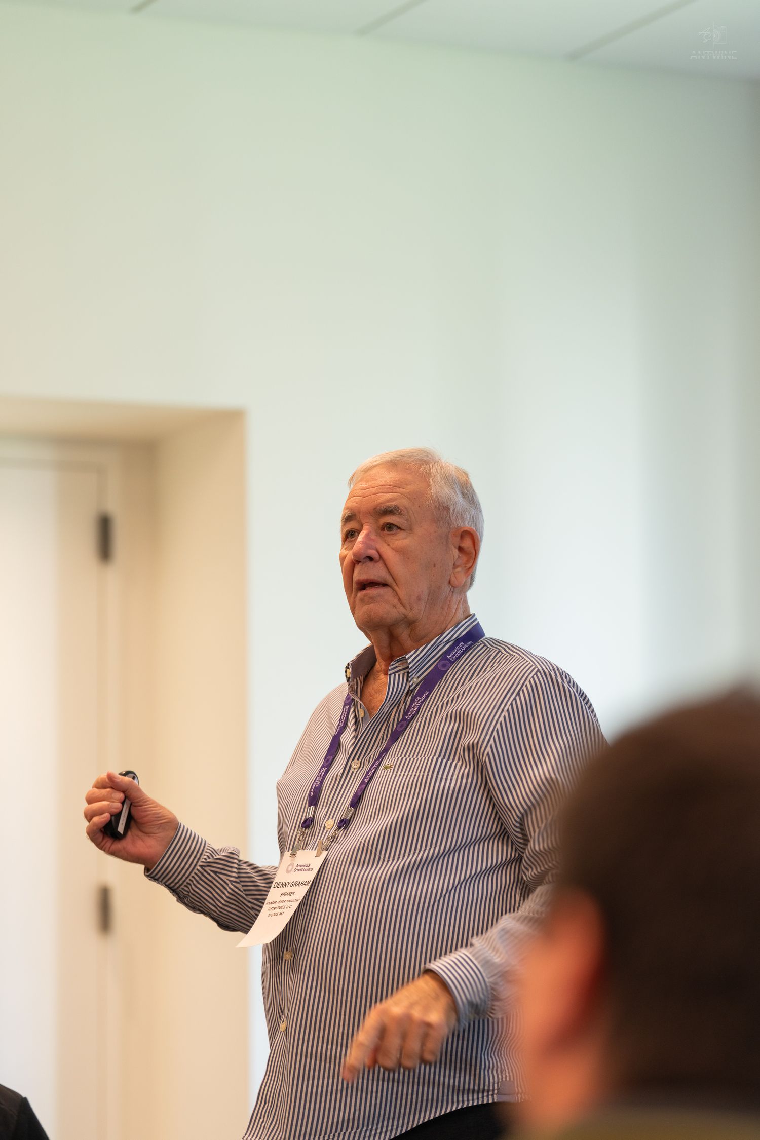 Elderly man in striped shirt presenting at a conference, holding a clicker while gesturing to an audience.