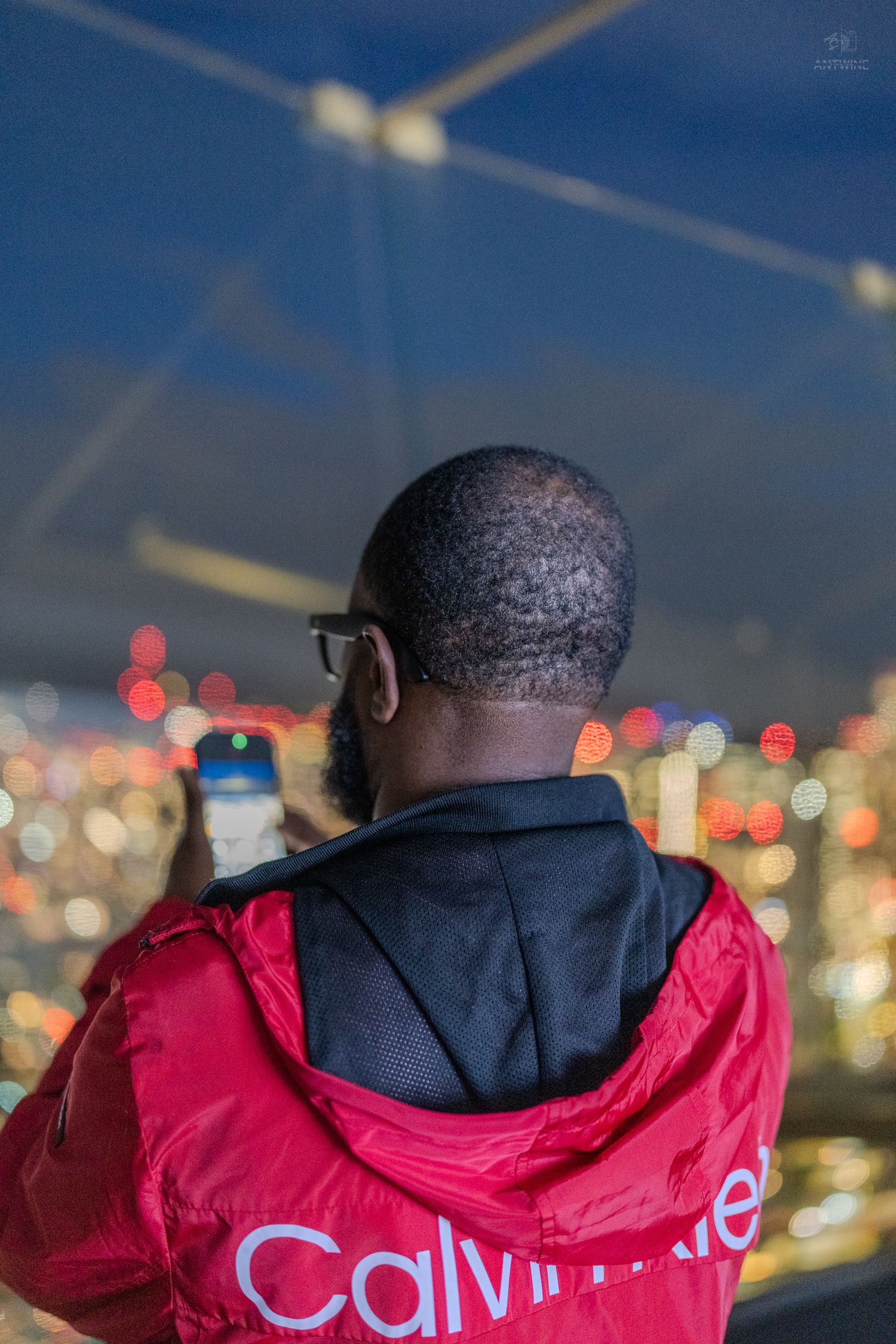 A man in a red Calvin Klein jacket photographs a bokeh city skyline at night.