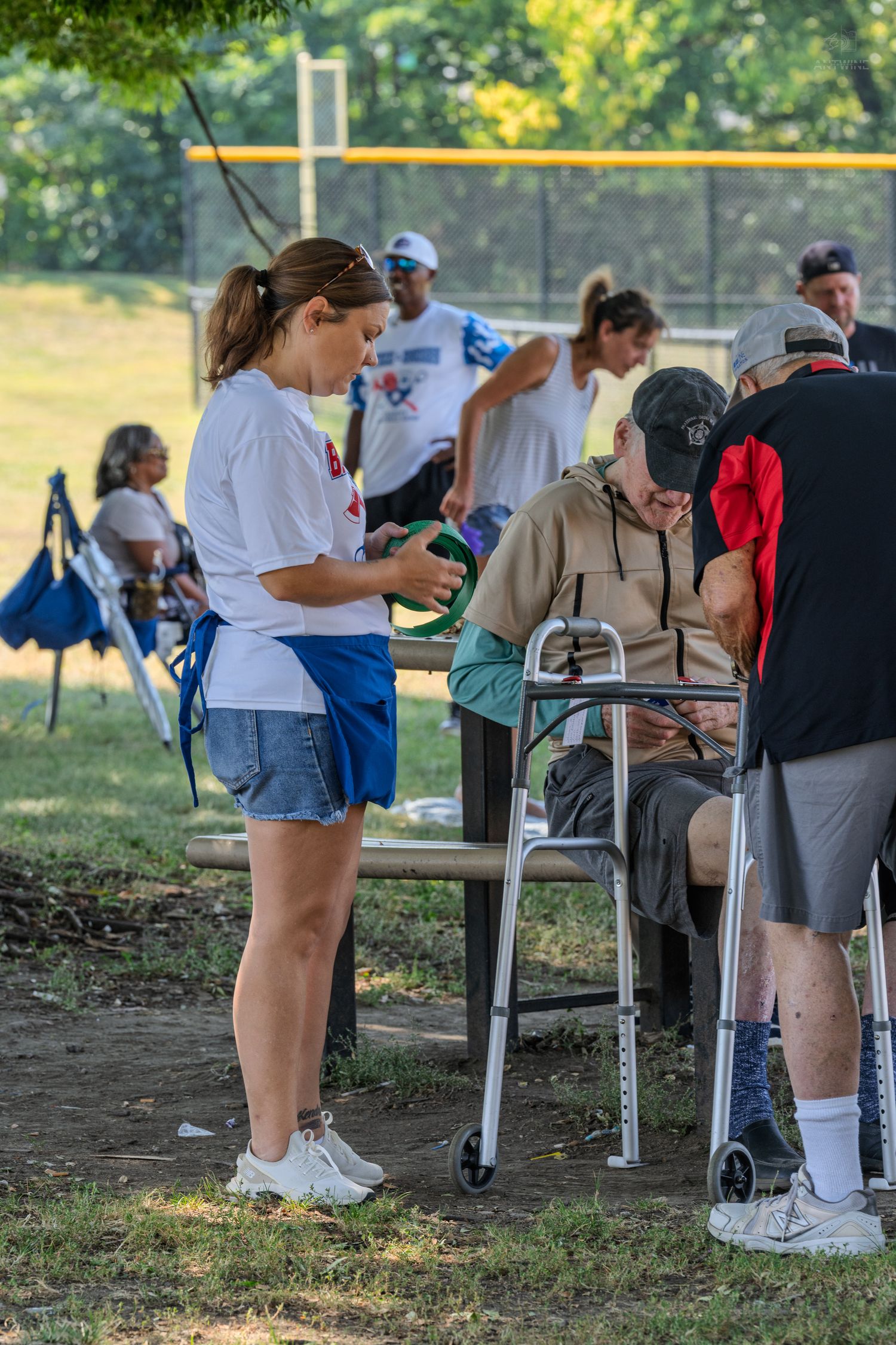 Woman in white shirt assists elderly man with walker at outdoor park picnic table during community event.