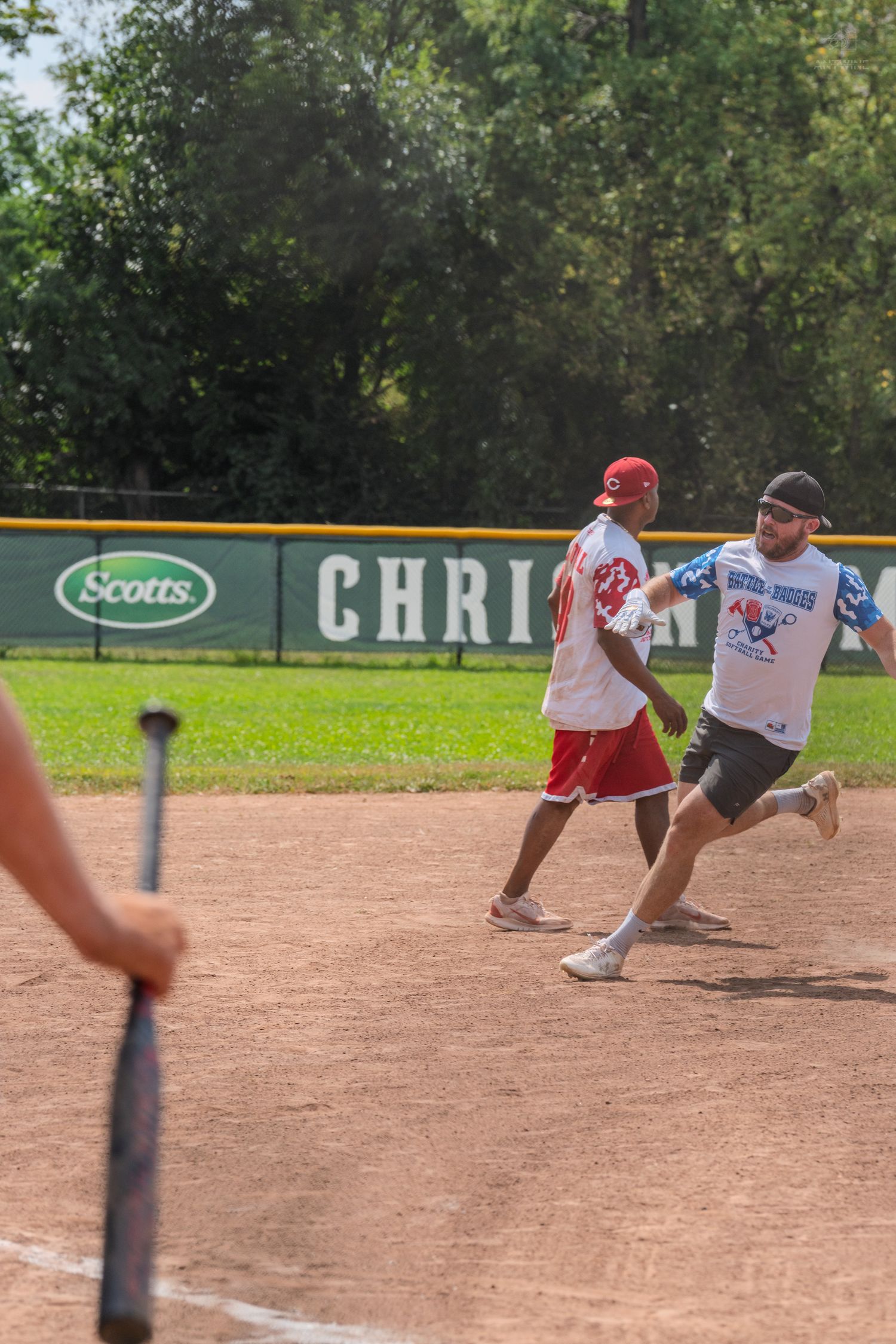 Players running bases during a sunny outdoor softball game with a Scotts sponsor banner visible in the background.