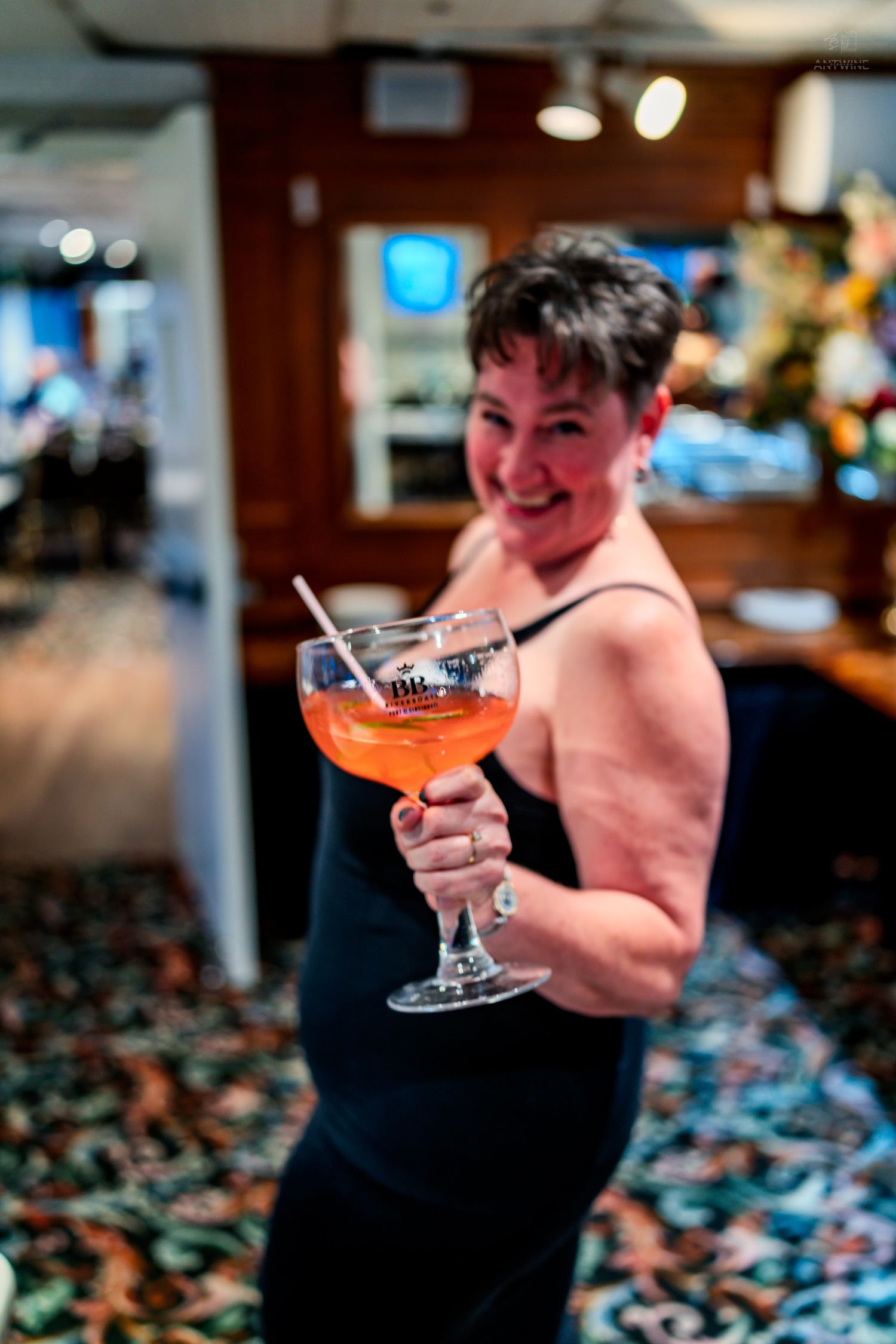 Woman in black dress smiling and holding a large cocktail glass with an orange drink in a bar setting.