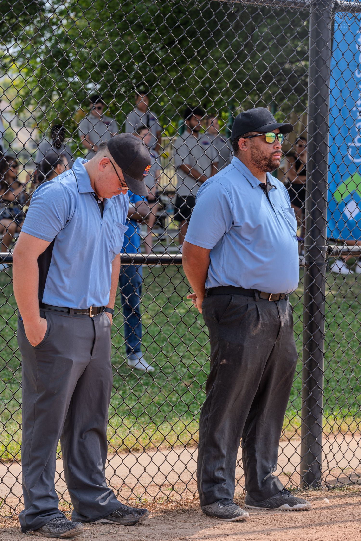 Two umpires in light blue shirts and black pants standing near a chain-link fence at a softball game.
