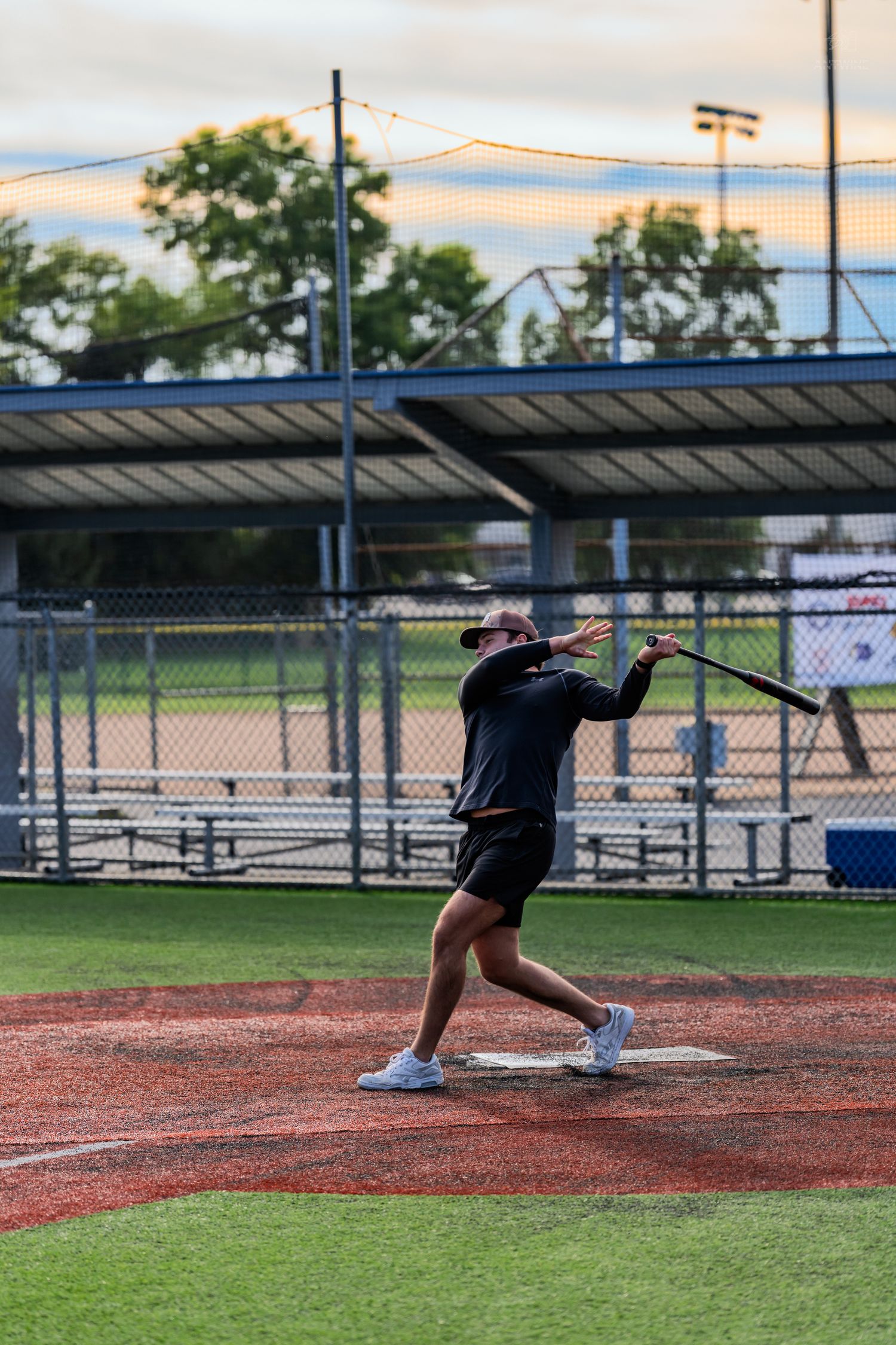 A baseball player in black athletic wear swings a bat at home plate on a well-maintained field at dusk.