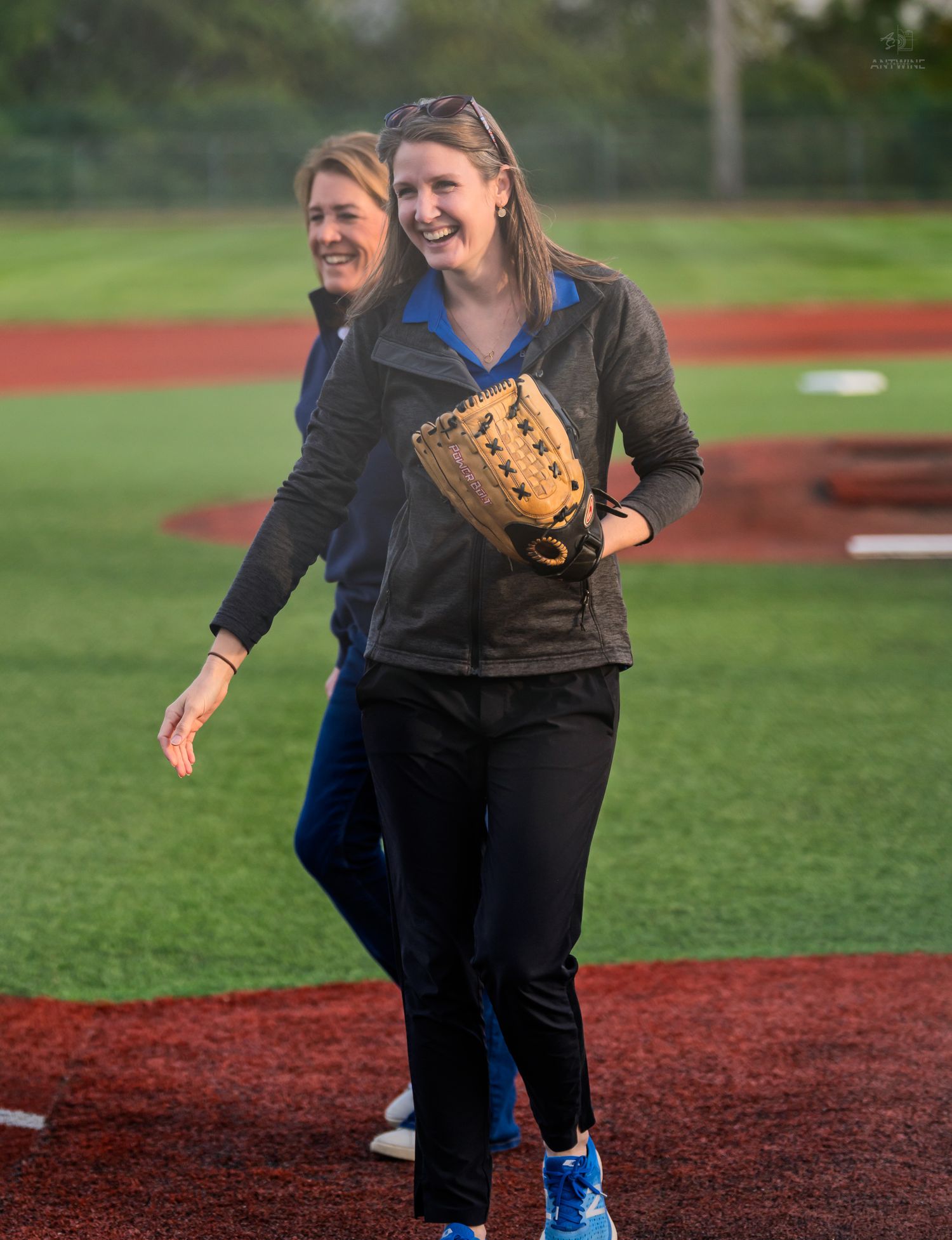 Smiling woman wearing a baseball glove walks off the field at an outdoor softball game.