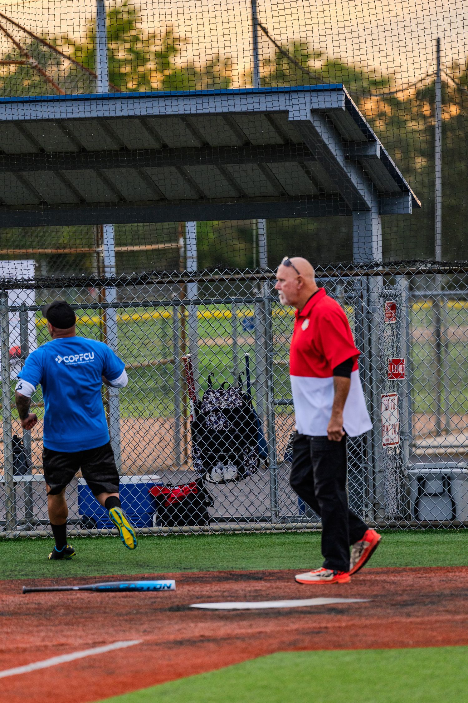 Two men on a baseball field, one in blue shirt running, another in red polo watching near home plate.