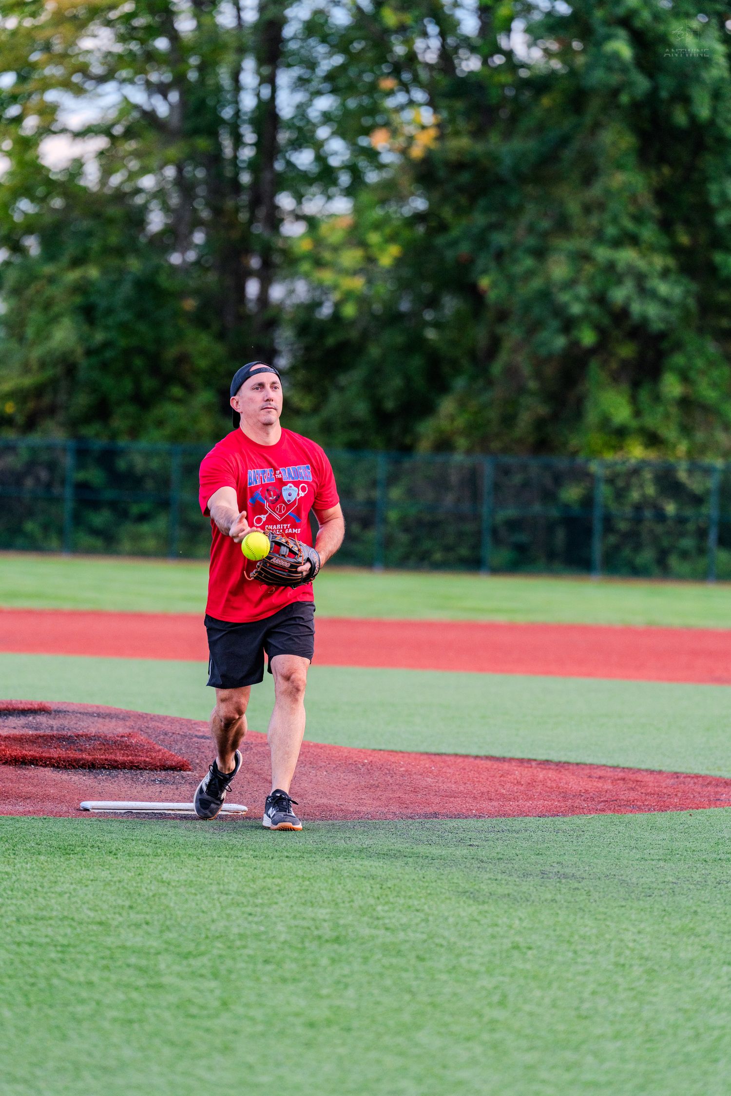 A man in a red shirt pitches a yellow softball on a red dirt infield during an outdoor game.