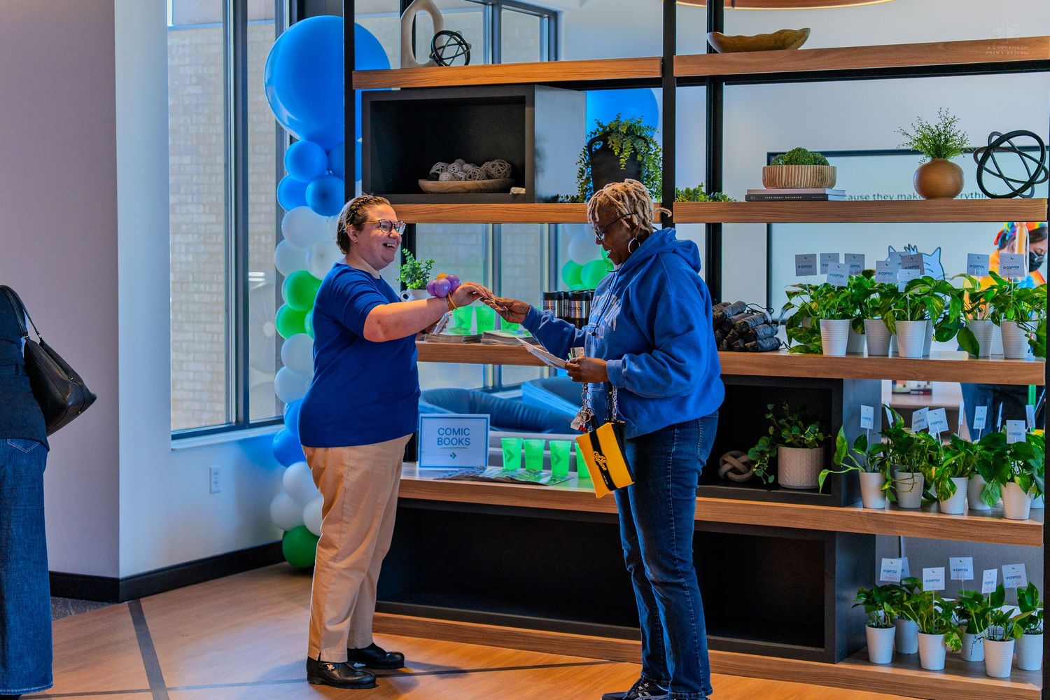 Two women exchanging items in a modern retail space decorated with blue balloons and green plants on shelves.