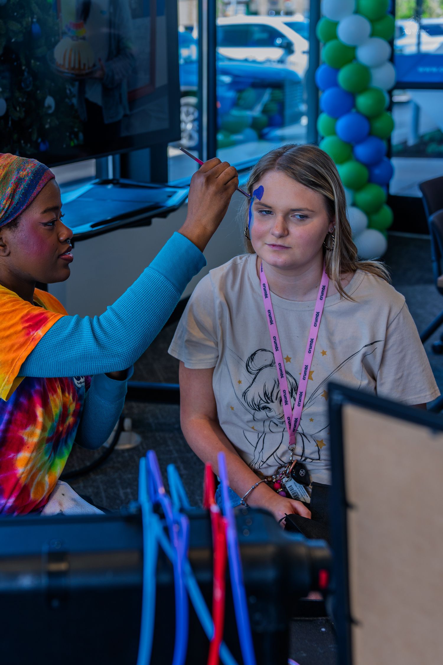A face painter applies blue paint to a young girl's forehead at a colorful event with balloon decorations.