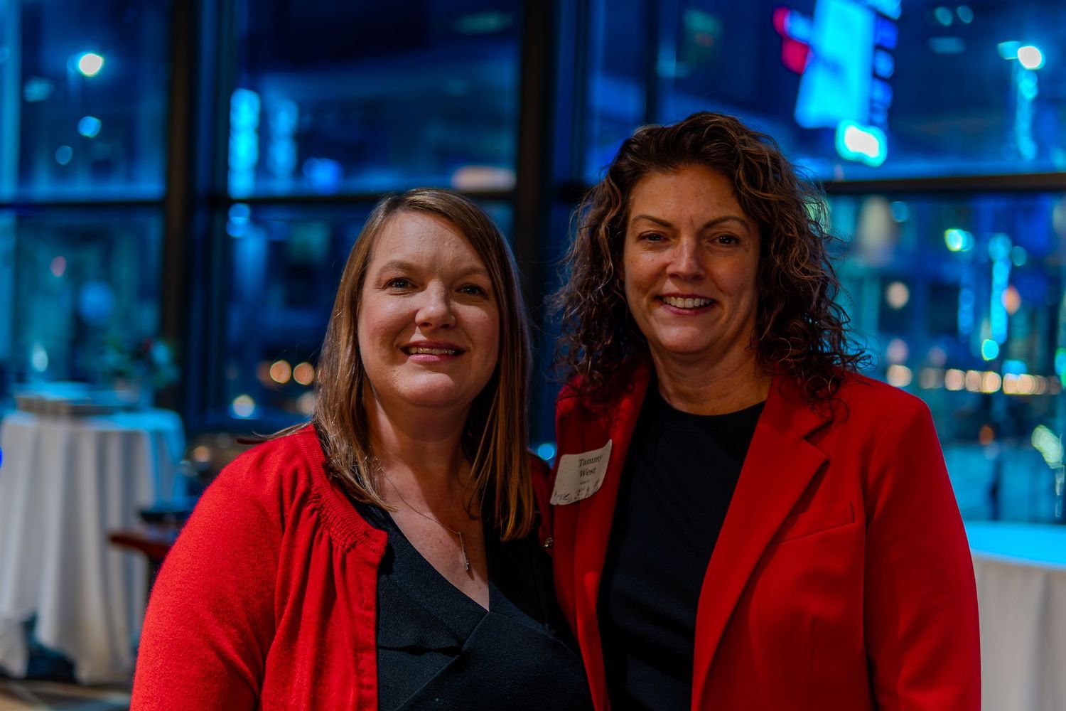 Two women in red blazers smile together at an evening corporate event with city lights in the background.