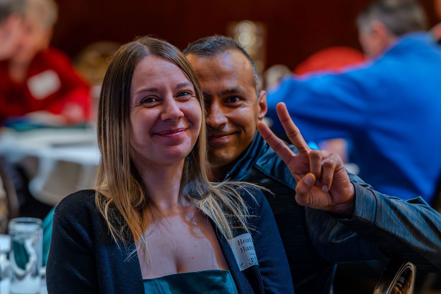 Two smiling people pose for a photo at an event, with a man making a peace sign gesture.