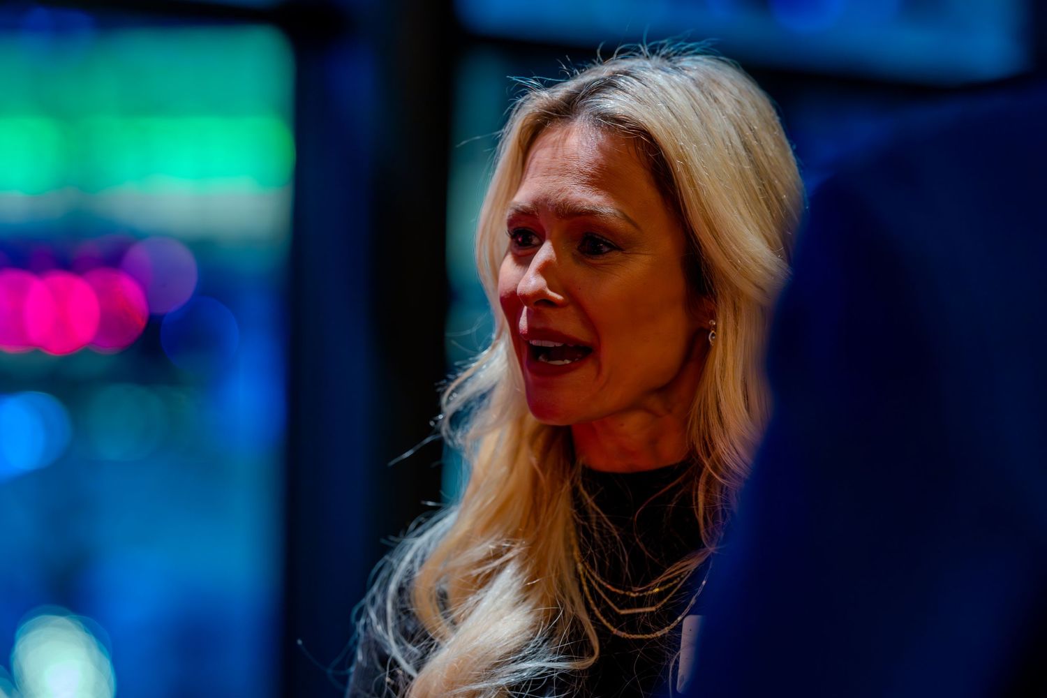 Blonde woman speaking at a dimly lit event with colorful bokeh lights in the background.