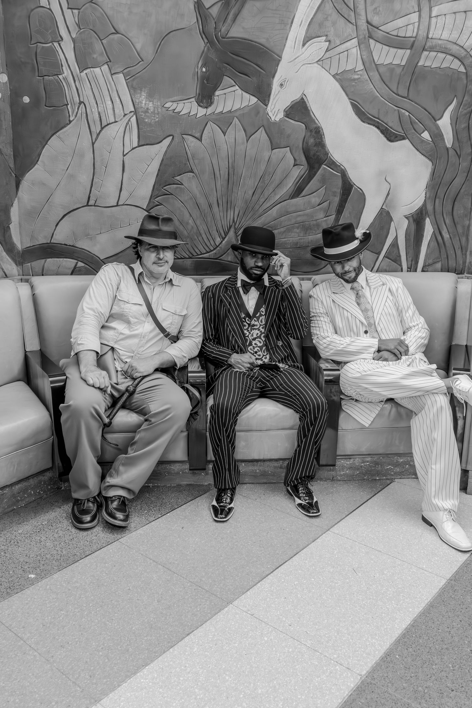 Three stylish men in suits and fedora hats sit on a bench in front of an Art Deco mural, black and white photo.