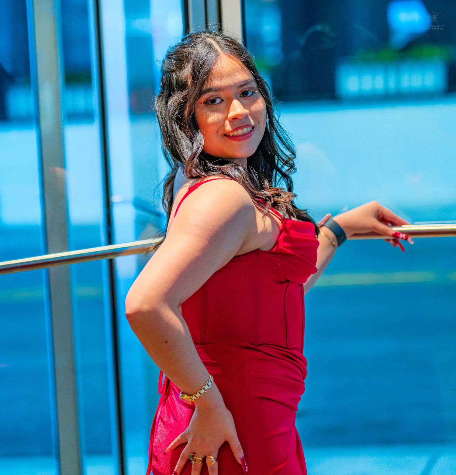 Young woman in a red formal gown posing gracefully by a glass railing at a prom venue.