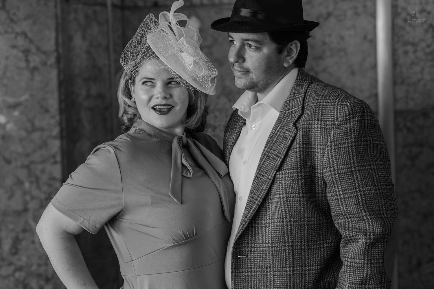 Black and white photo of a couple in vintage 1940s attire, woman in fascinator hat and man in fedora and plaid suit.