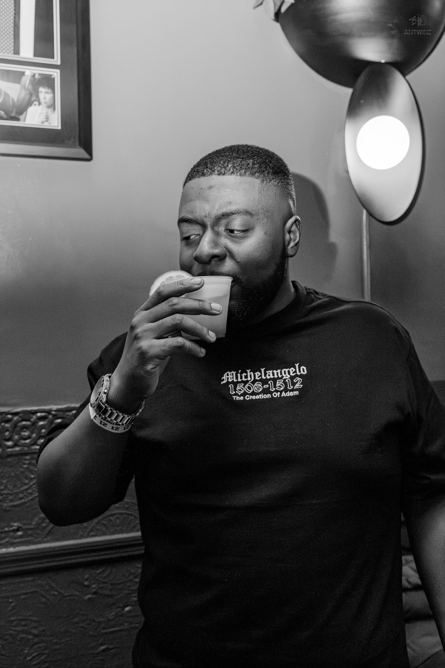 Black man in Michelangelo graphic tee sipping from a cup under studio light in black and white photo.