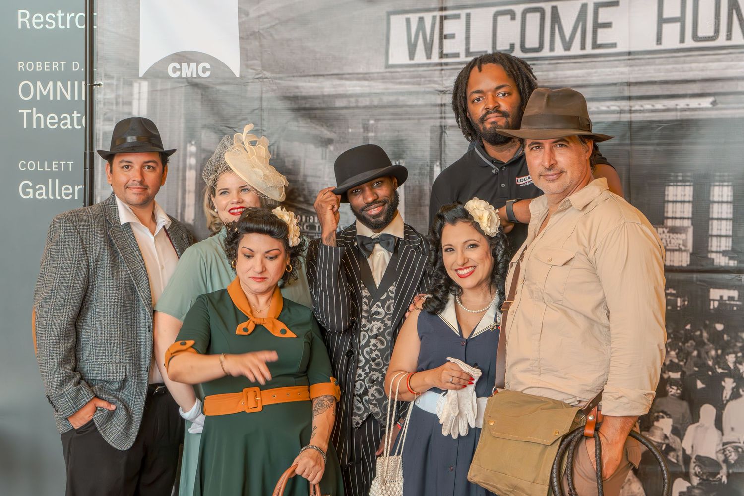 Group of seven people in 1940s vintage costumes posing in front of a Welcome Home theater backdrop.
