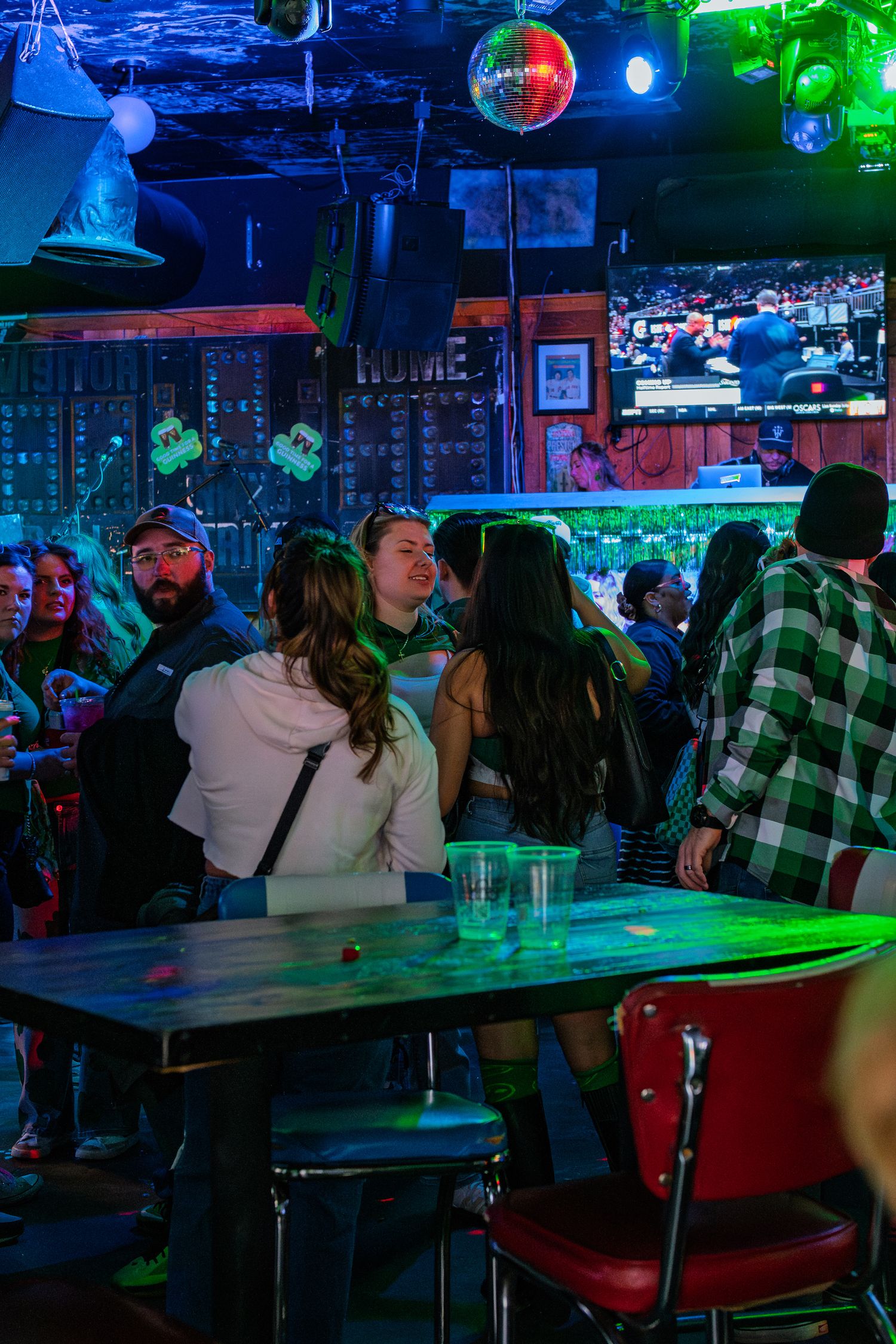 Crowded bar with neon green lights, disco ball, and patrons socializing near a table with drinks.