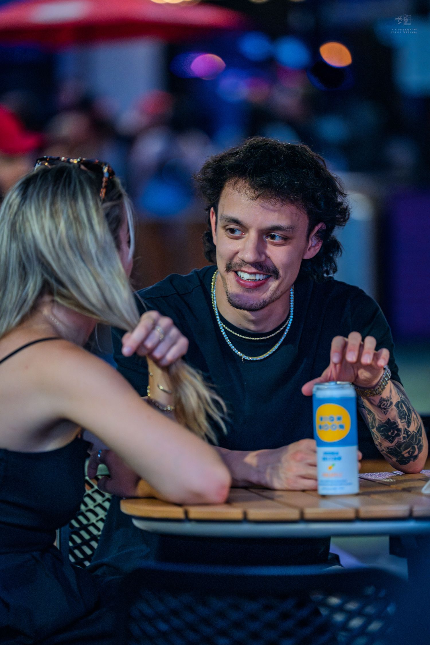 A young man with tattoos and a chain necklace smiles at a blonde woman at an outdoor table with a canned drink.