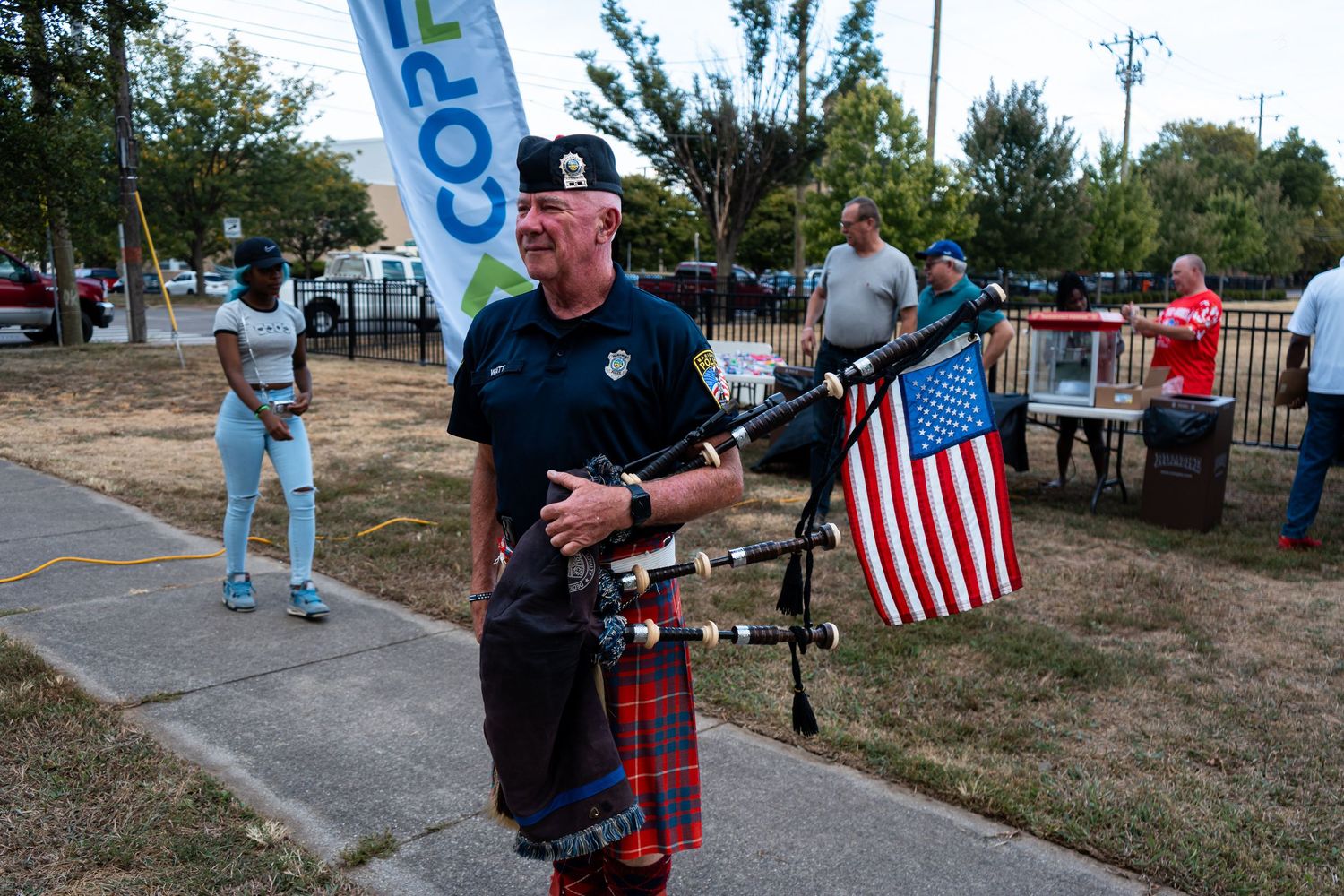 Man in traditional Scottish kilt plays bagpipes while holding an American flag at an outdoor event.