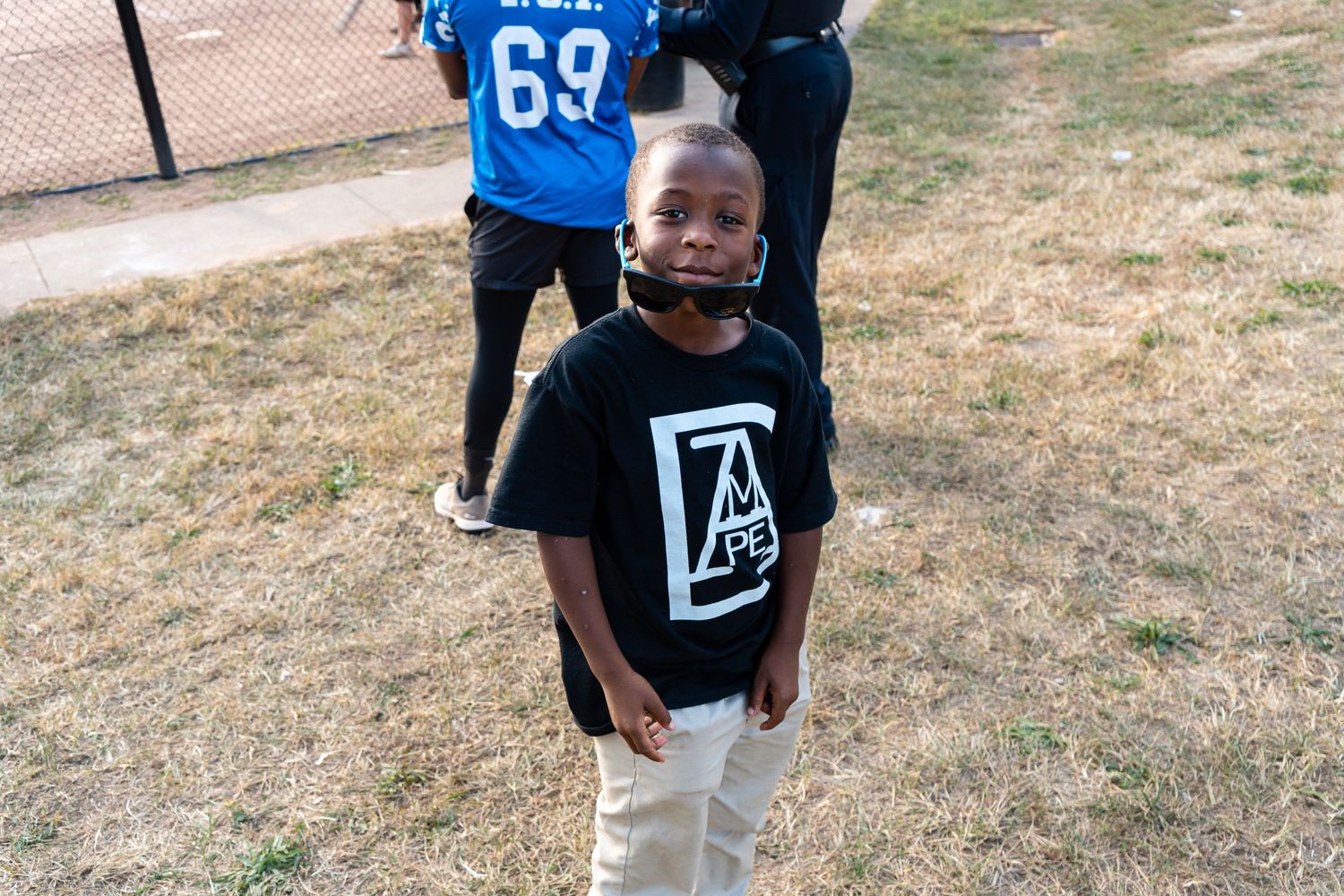 Young child smiles wearing a black graphic t-shirt while standing on a grass field at a sports event.