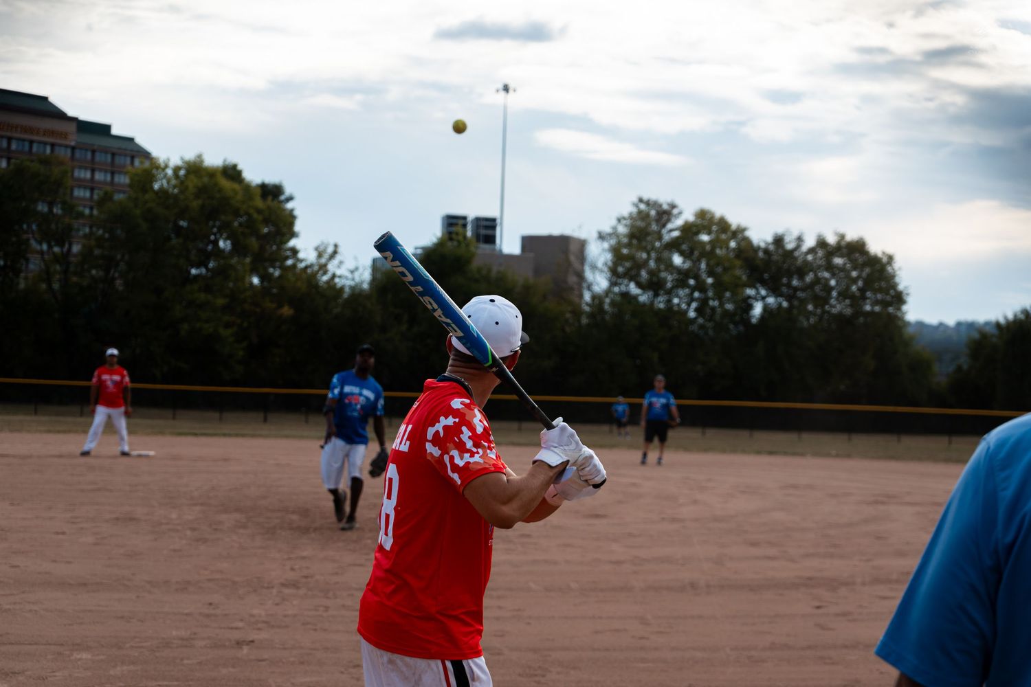 A batter in a red jersey swings at a softball as the ball approaches on an outdoor dirt field.