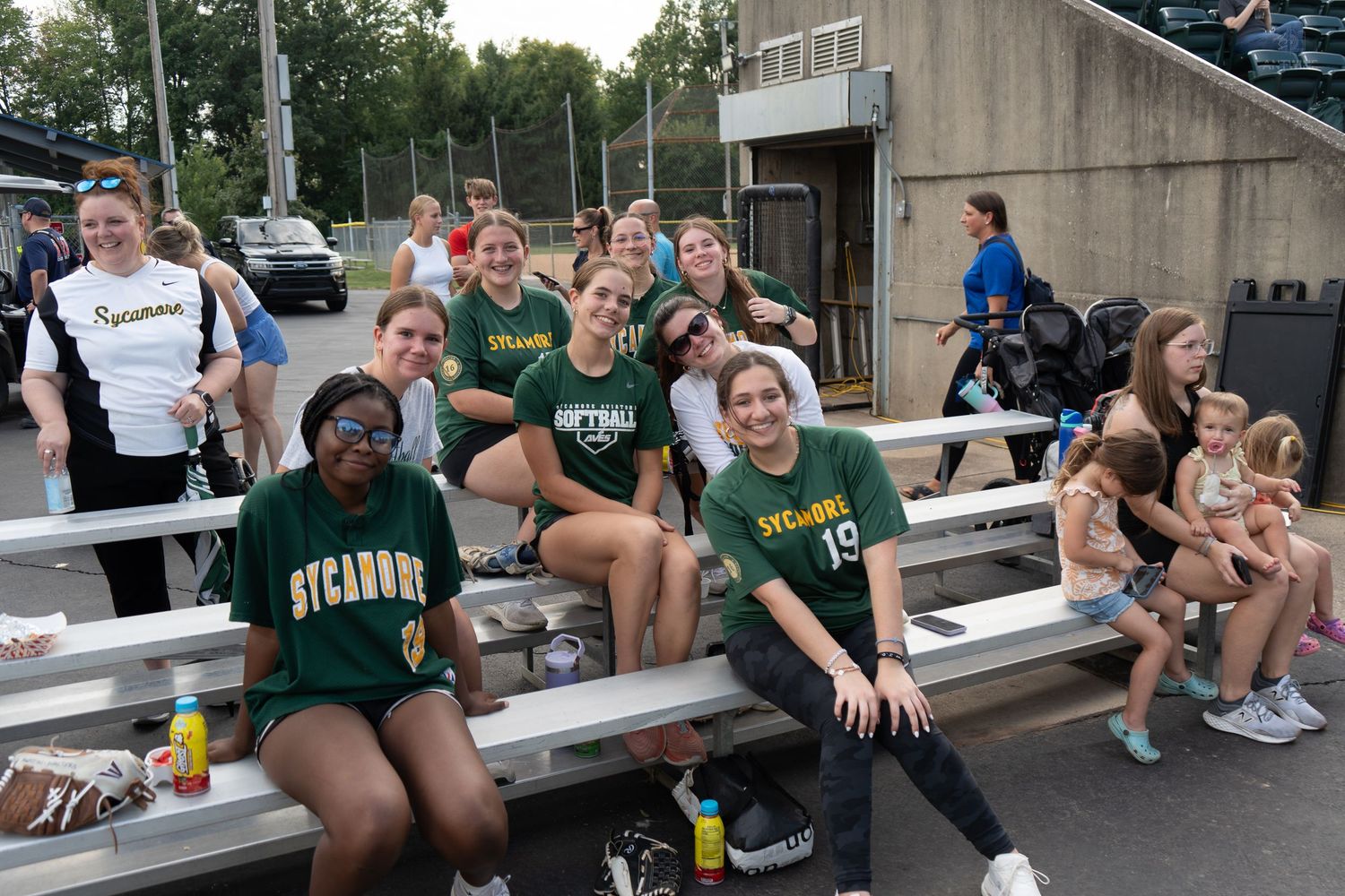 Sycamores softball team posing together on bleachers after a game, smiling in green and gold uniforms.