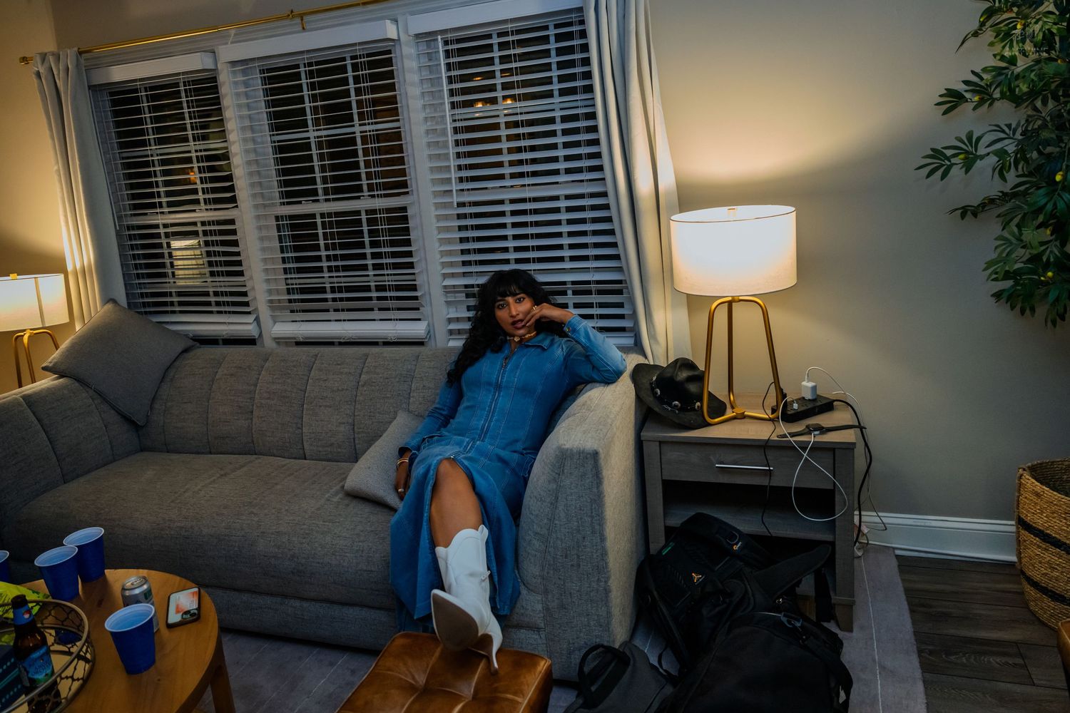Woman in blue dress relaxing on gray couch with white boots in cozy living room with lamp and shuttered windows.