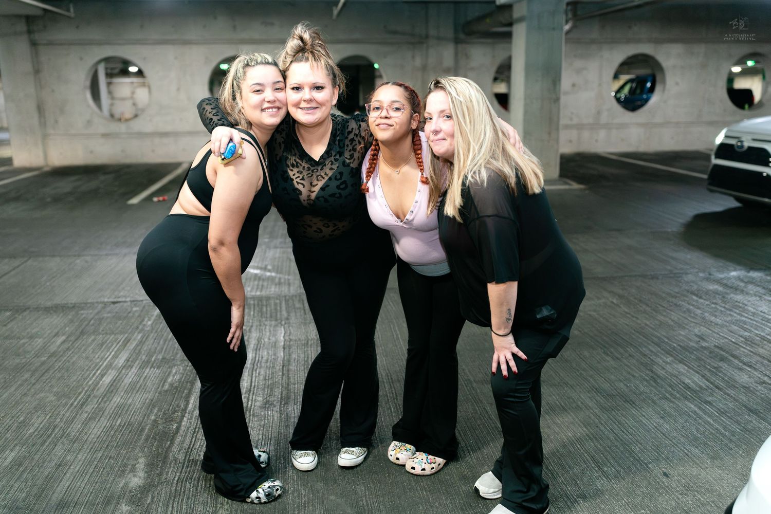 Four women pose together in a parking garage wearing black outfits for a night out.
