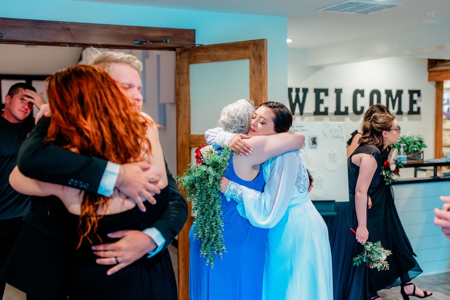 Wedding guests exchanging warm hugs near a Welcome sign at a rustic venue, with a bride in blue holding greenery.