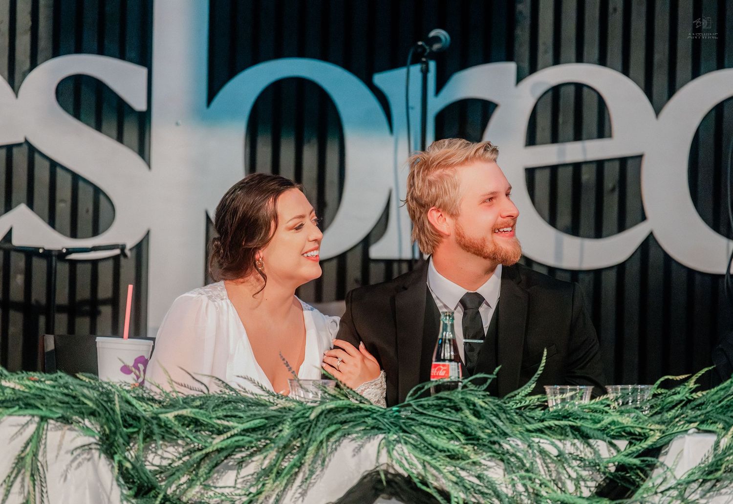 Smiling couple sits at decorated head table with greenery at formal event, woman in white, man in black suit.