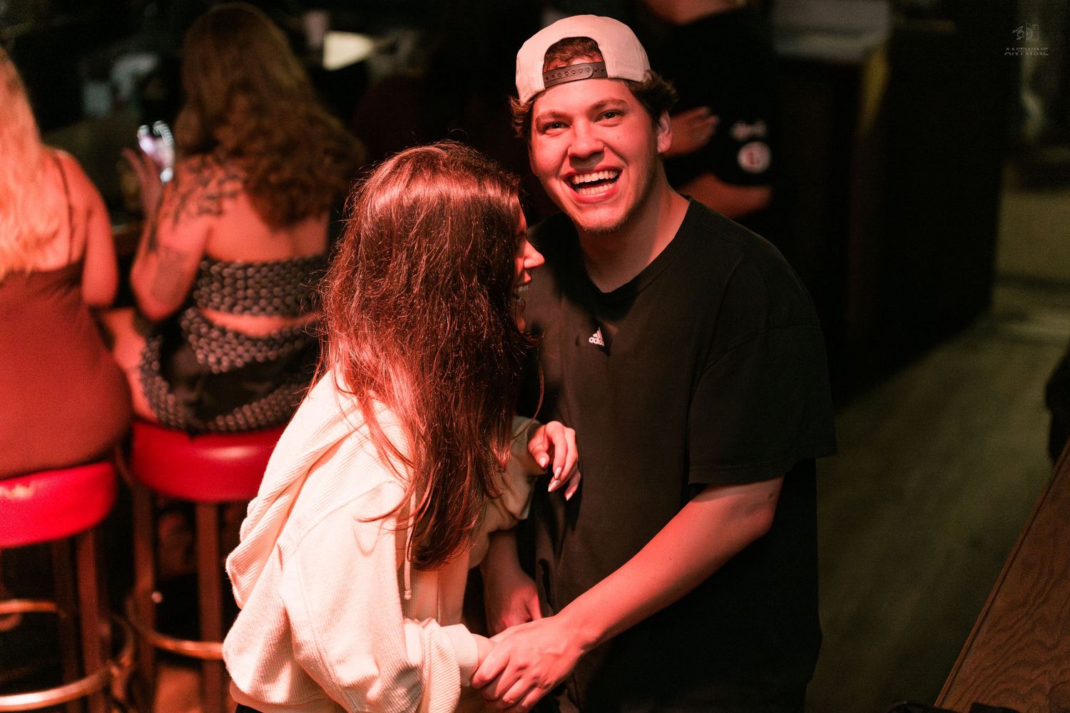 A smiling young man in a backwards cap laughs with a woman at a dimly lit bar or nightclub.