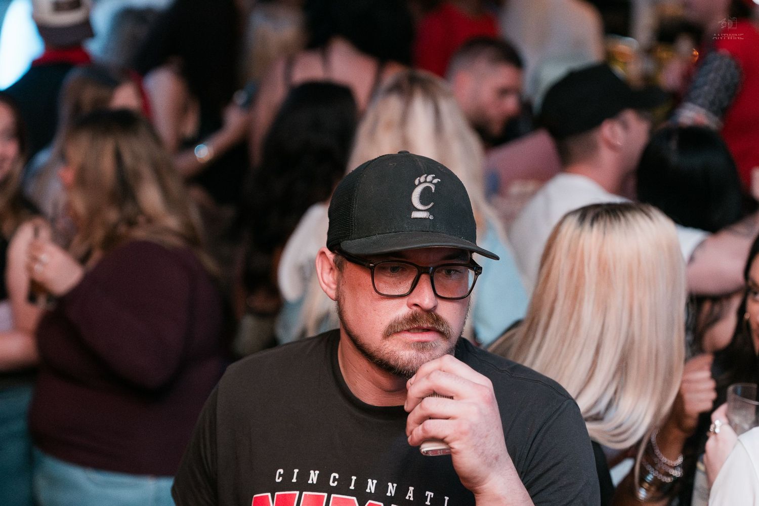 A man in a Cincinnati hat pensively touches his chin at a crowded bar event.