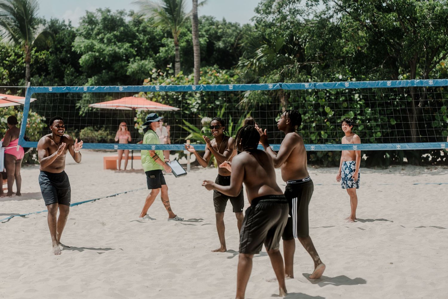 Group of people playing beach volleyball on white sand surrounded by lush tropical greenery and umbrellas.