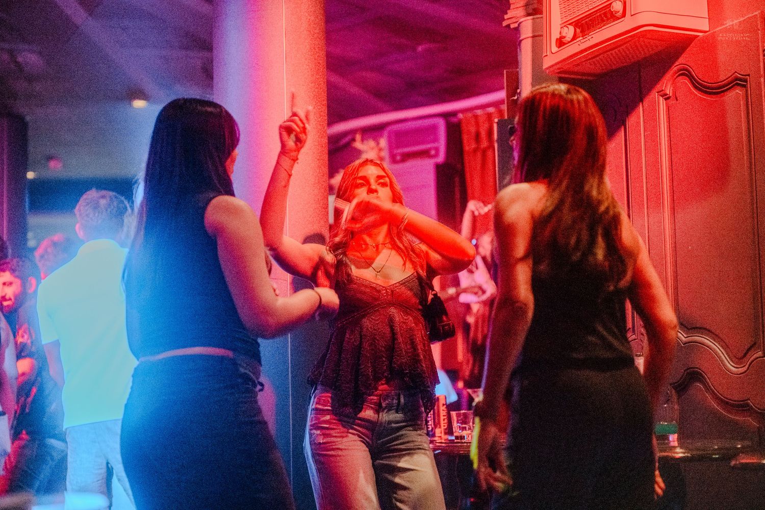 Three women dance and celebrate under vibrant red lights at a nightclub party.