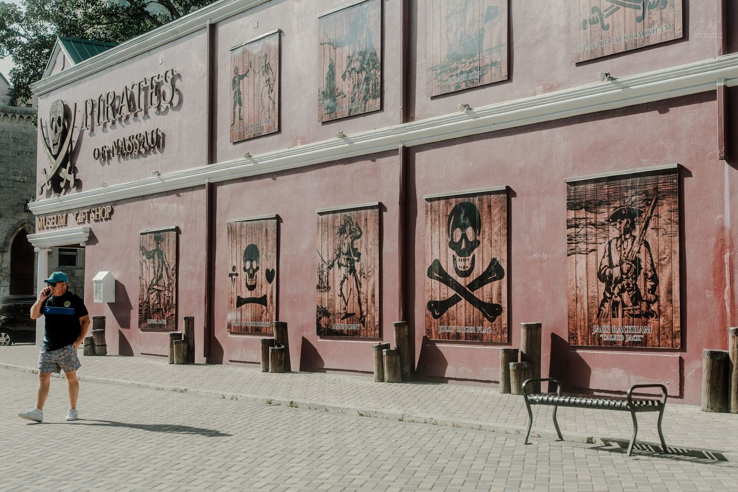 Tourist walking past a pirate-themed attraction with skull and crossbones artwork on a pink wall.